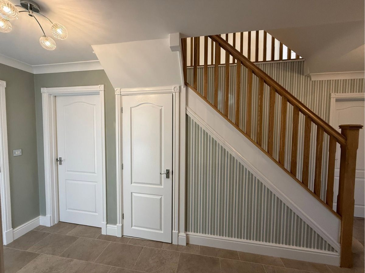 Decorated entrance hallway featuring soft green walls, white interior doors, striped wallpaper and a natural wood staircase.